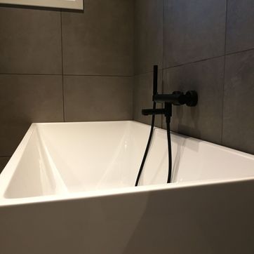 Moderne hvitt badekar med mørke fliser på vegg og sort batteri